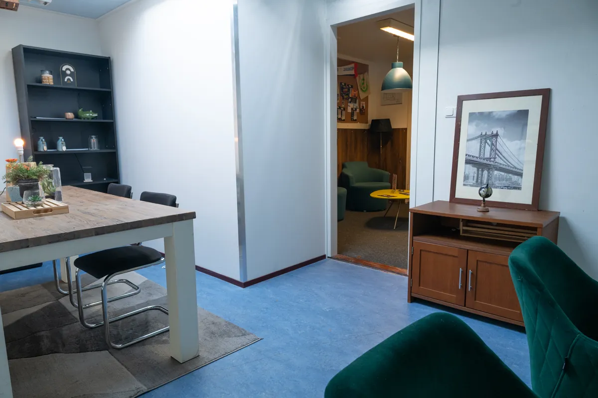 Een houten werktafel met stoelen, een groene fauteuil en kunst aan de muur in de co-workingruimte bij de entree