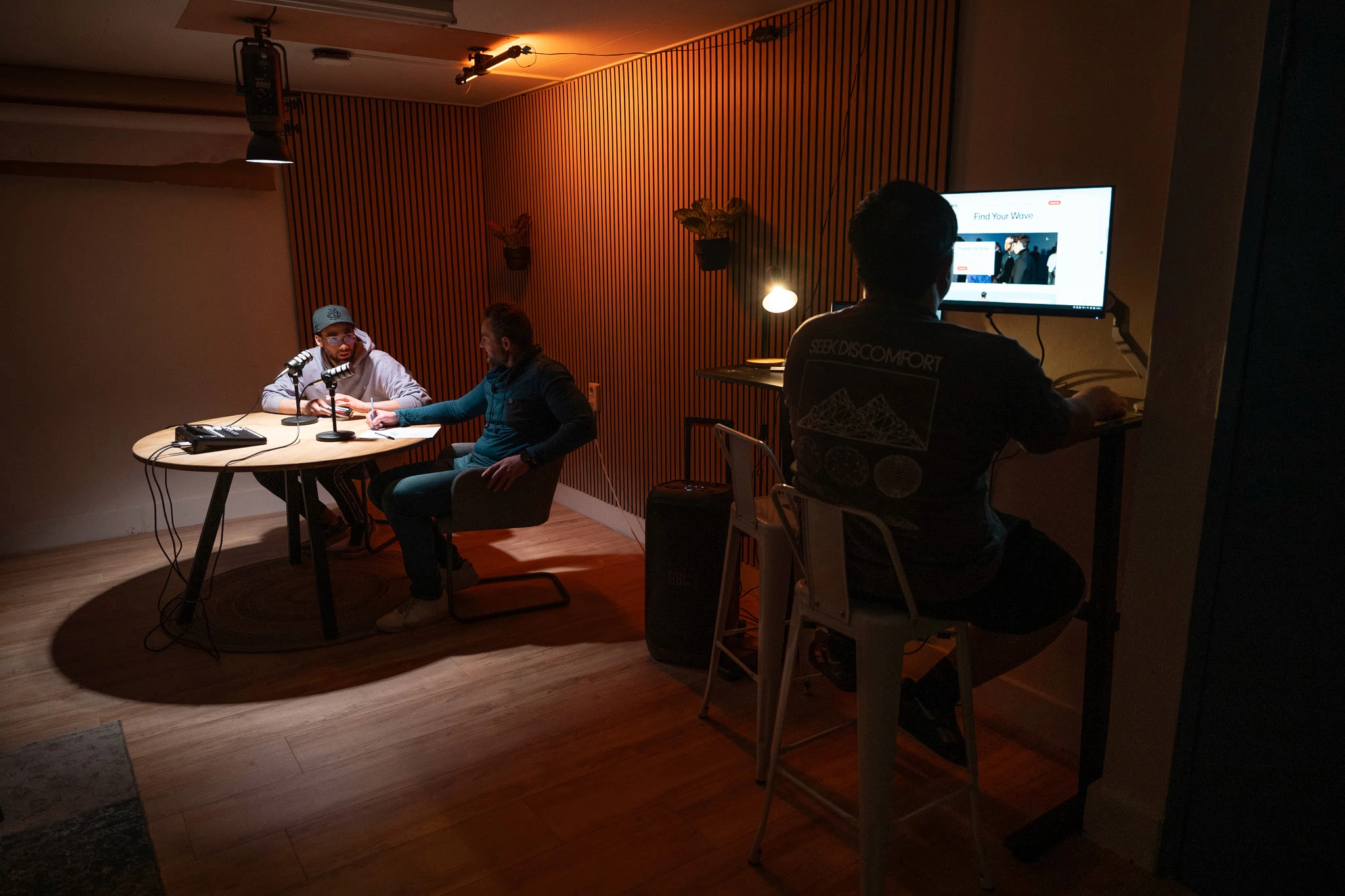 Een podcastopname aan tafel in de mediastudio, terwijl iemand anders aan het bureau werkt