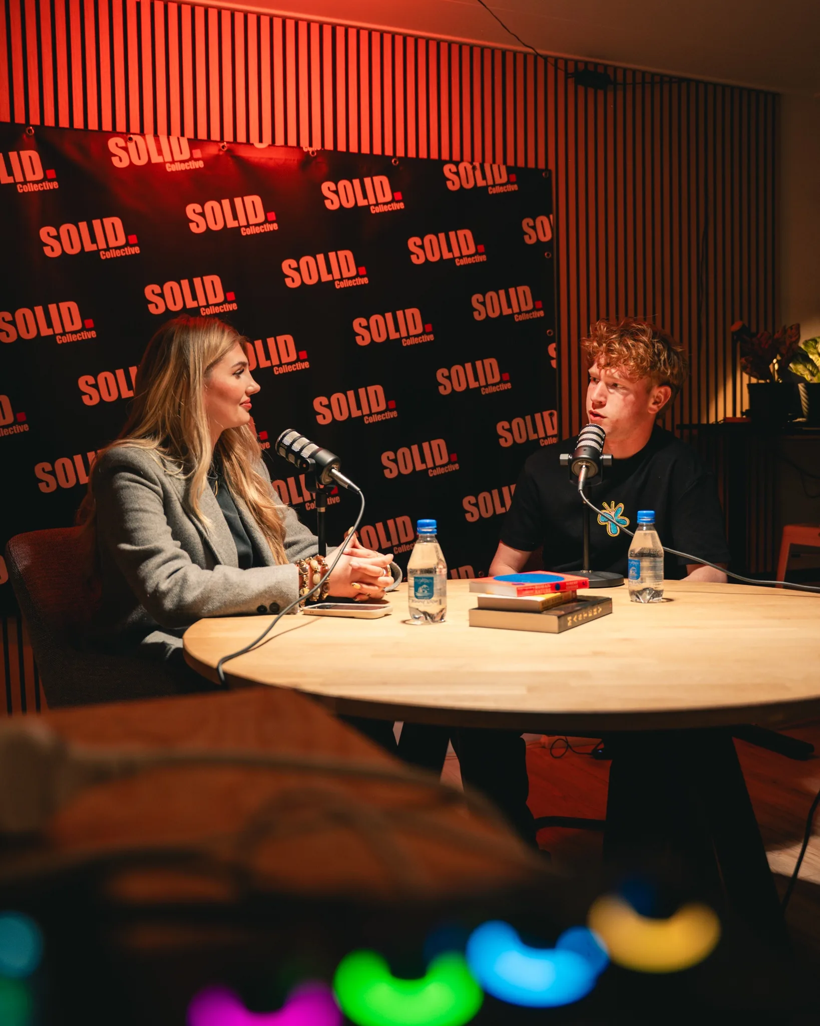 Twee mensen in gesprek aan een ronde tafel met podcastmicrofoons, voor een SOLID-backdrop in rood licht