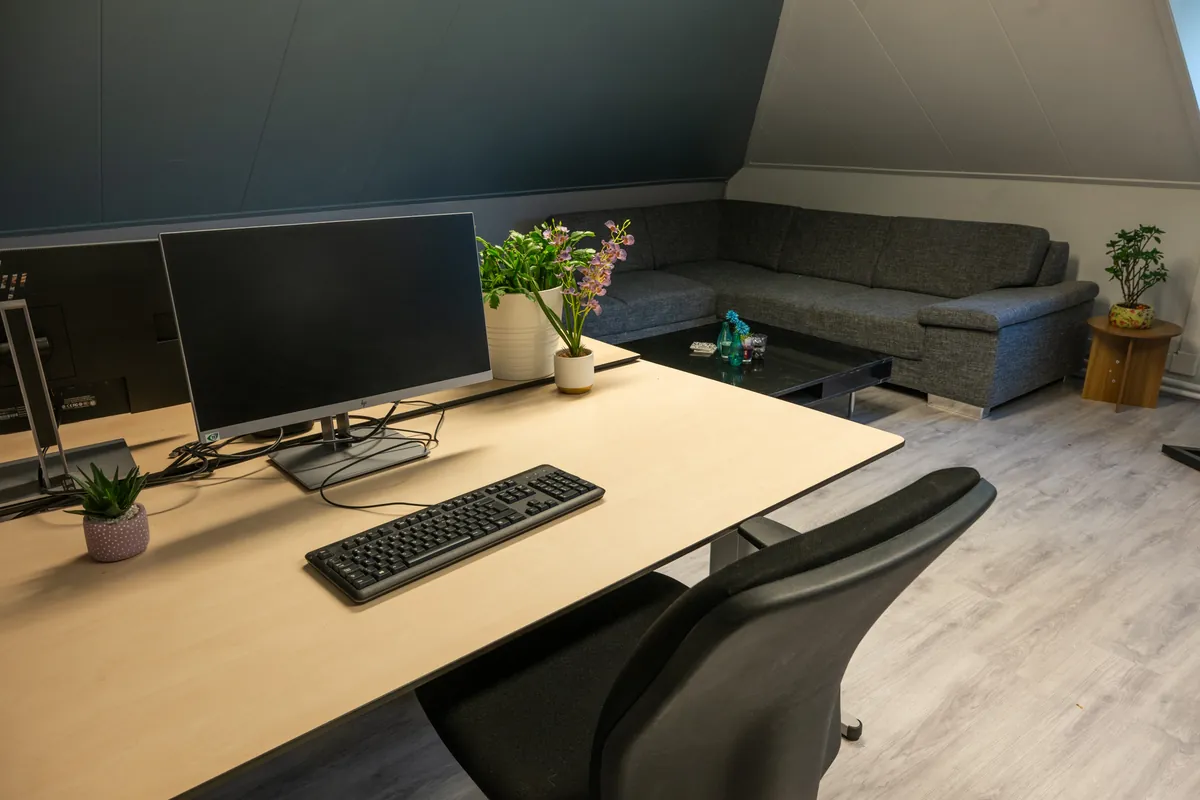 Een bureau met een groot scherm en een plant, met de hoekbank op de achtergrond, in de zolderkamer